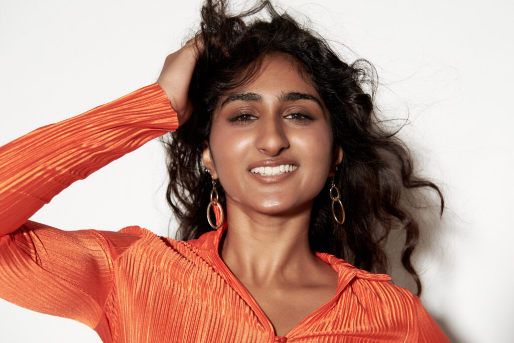 A headshot image of a female model with long, wavy, dark hair wearing an orange shirt against a white backdrop.