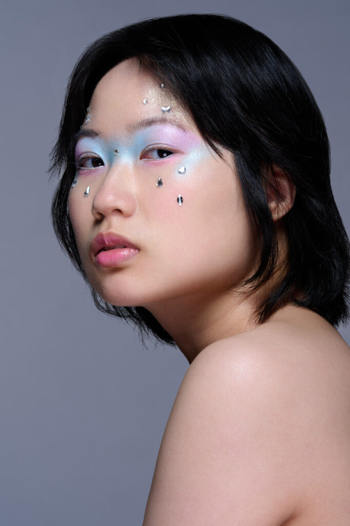 Model with purple, and blue makeup around the eyes and tear-drop shaped jewels on the skin of her face