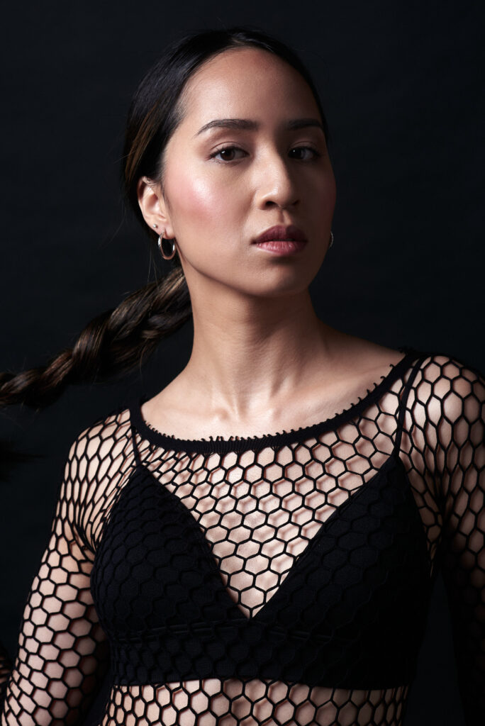 Model with braided hair in the studio wearing a net shirt against a black background.