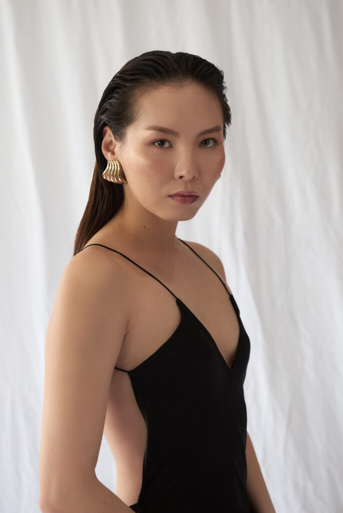 A model in an elegant black evening dress with thin straps against a white, fabric background.  She is wearing gold earrings.
