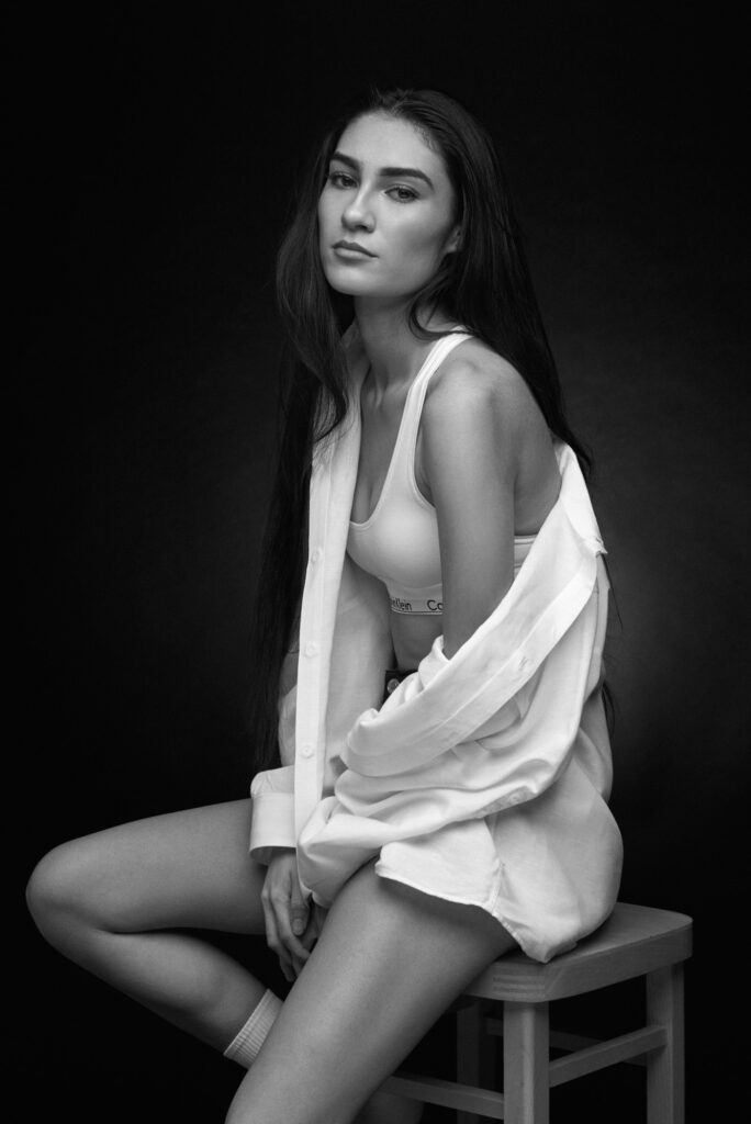 Black and white image of a model sitting in Calvins and an oversized white shirt on a stool in the studio